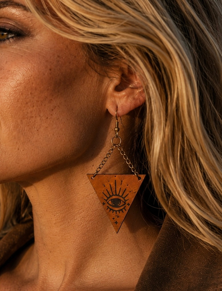 A close-up of a woman's ear wearing a dangling triangular wooden earring with a black engraved eye and sunburst design, set against warm golden lighting.